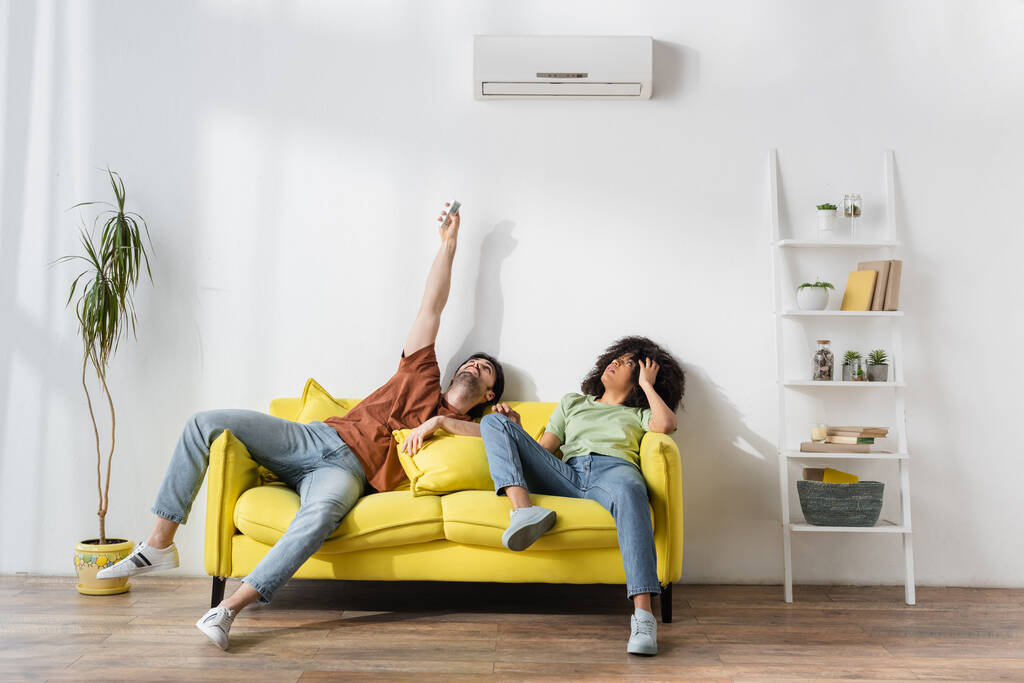 man met afstandsbediening in de buurt van moderne airconditioner en Afrikaanse Amerikaanse vriendin die last heeft van warmte - Foto, afbeelding