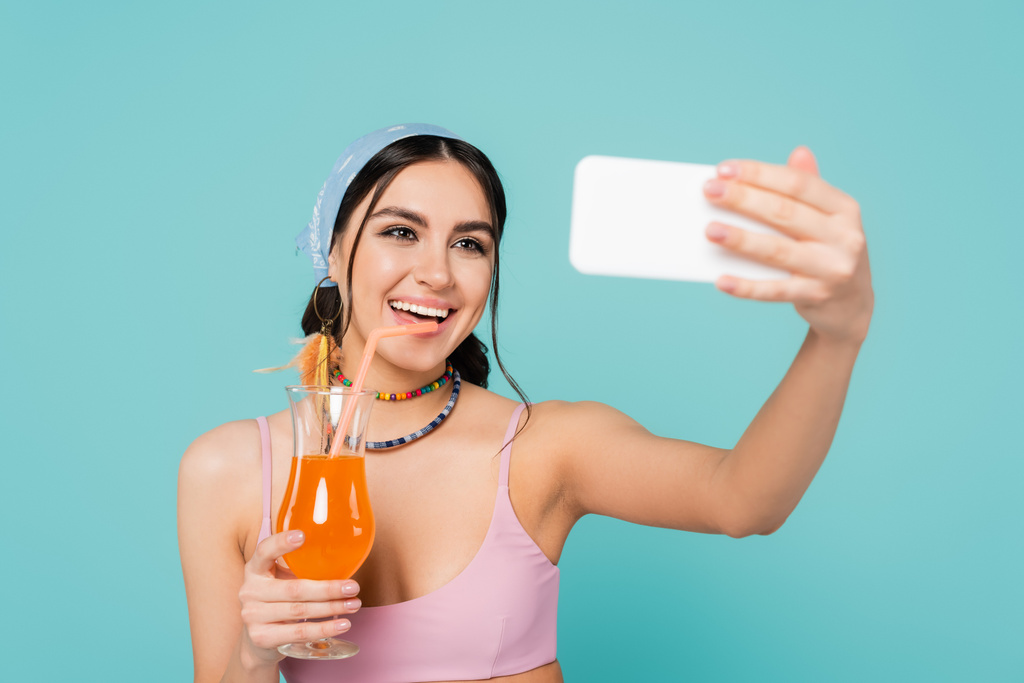 Mujer positiva con cóctel tomando selfie en un smartphone borroso aislado en azul  - Foto, imagen