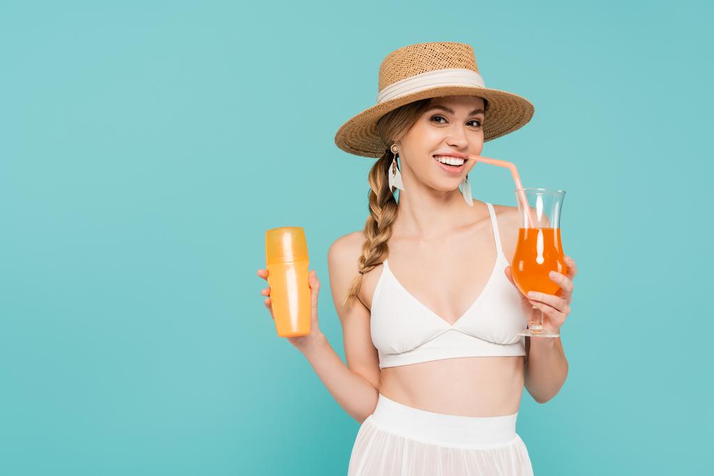 Smiling woman in straw hat holding sunscreen and cocktail isolated on blue  - Photo, Image