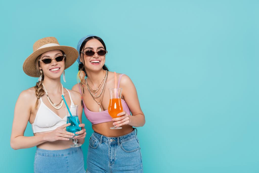Mujeres sonrientes en gafas de sol sosteniendo cócteles aislados en azul  - Foto, Imagen