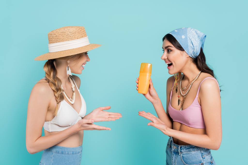 Smiling women in tops pointing at sunscreen isolated on blue  - Photo, Image