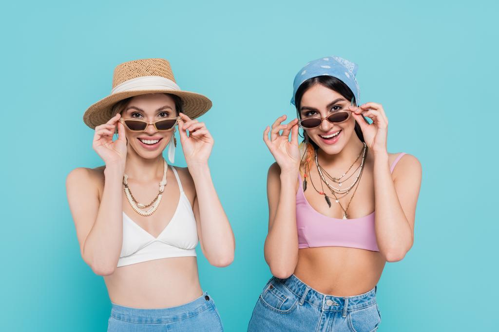 Положительные женщины в верхней части держат солнечные очки изолированы на синий  - Фото, изображение