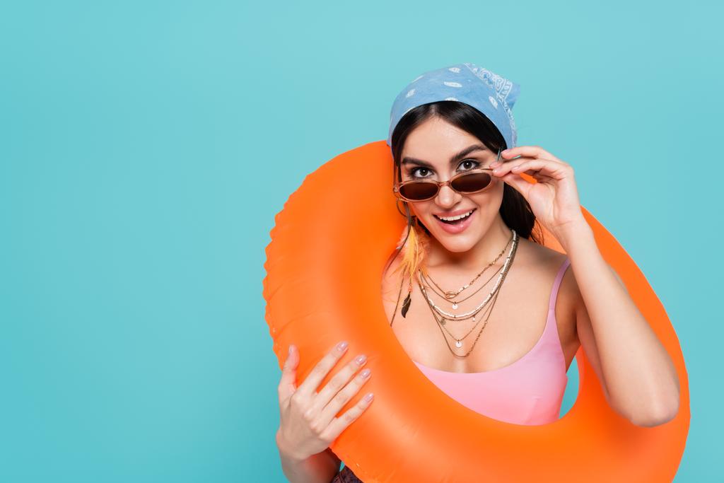 Веселая женщина в солнечных очках, держащая плавательное кольцо изолировано на голубом - Фото, изображение
