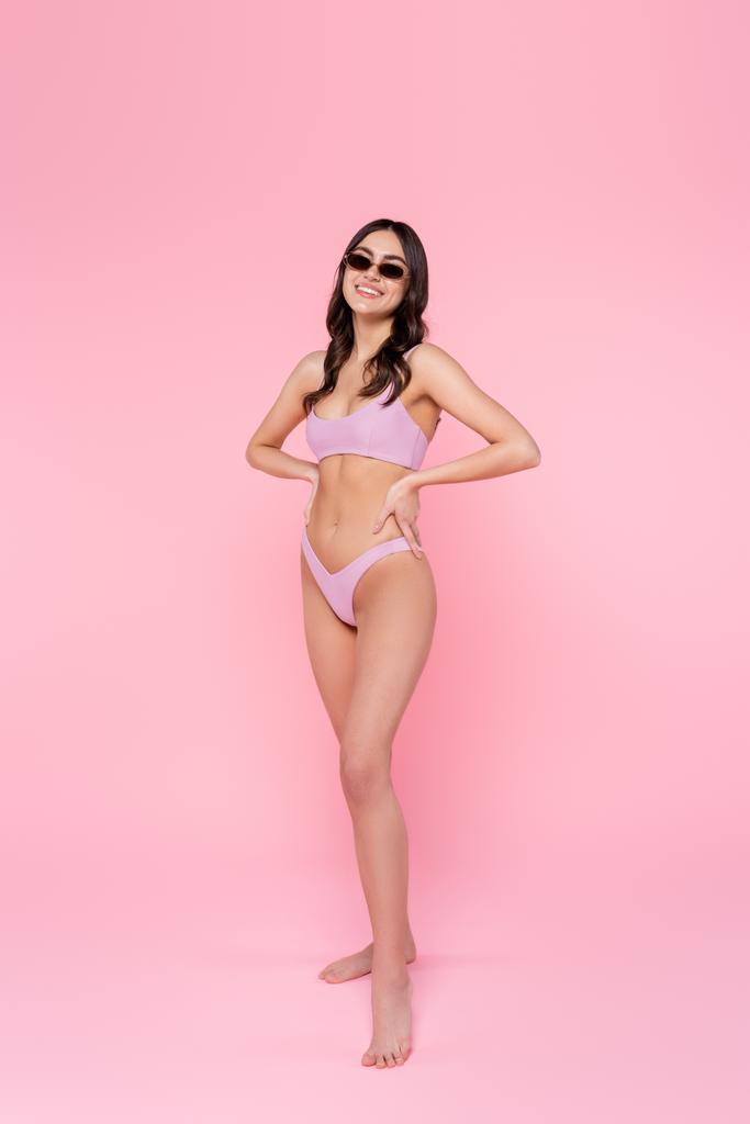Щаслива жінка в купальнику і сонцезахисних окулярах тримає руки на стегнах на рожевому фоні
  - Фото, зображення