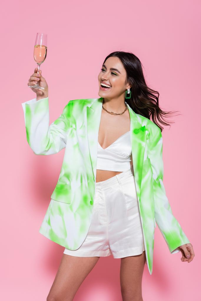 Positive woman in jacket holding glass of champagne on pink background  - Photo, Image