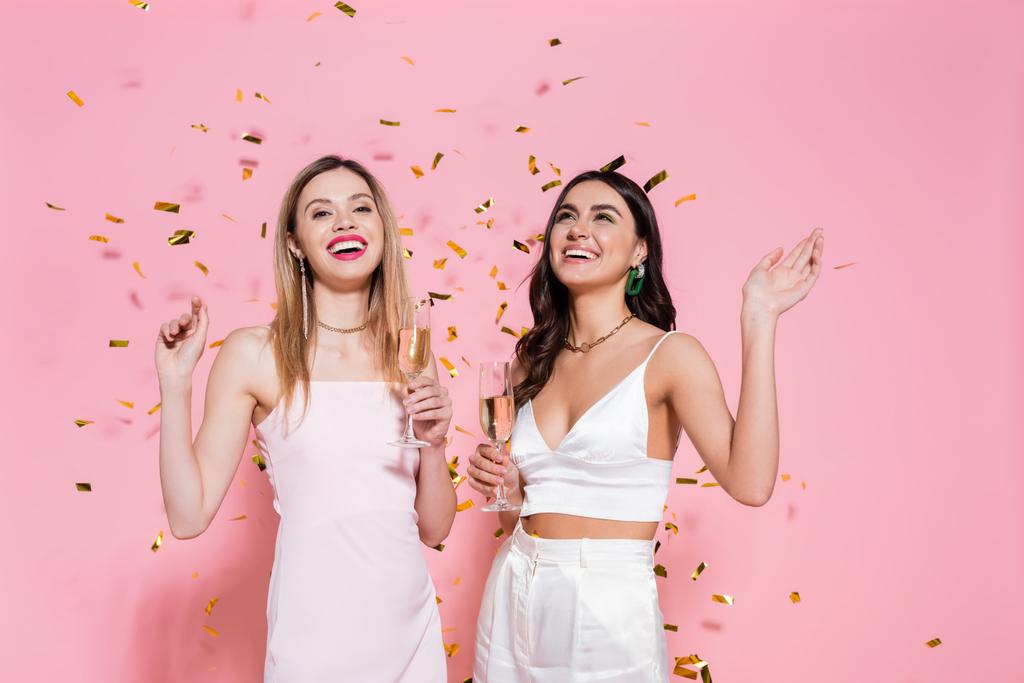 Positiivinen brunette nainen samppanjaa seisoo lähellä ystävä ja konfetti vaaleanpunainen tausta  - Valokuva, kuva