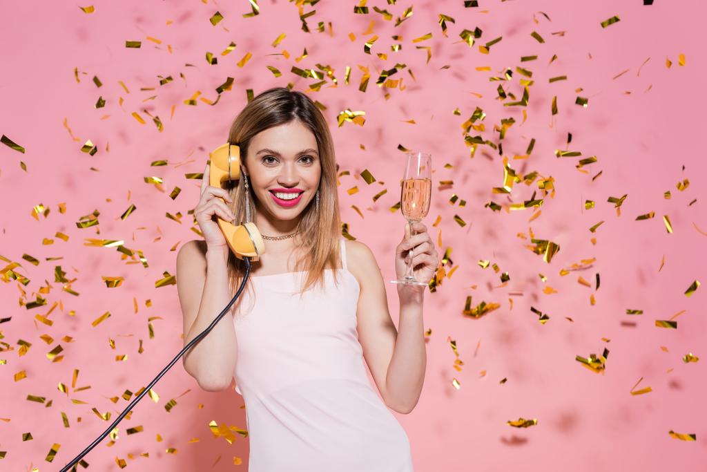 Cheerful woman with champagne talking on telephone under confetti on pink background  - Photo, Image