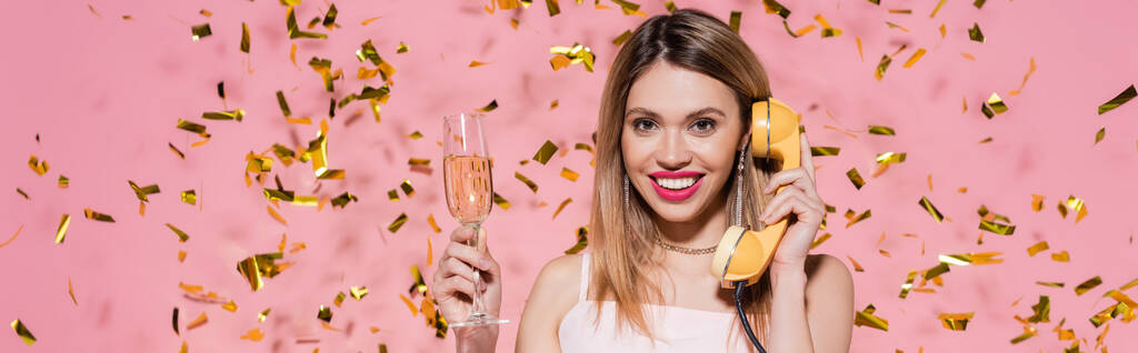 Mujer sonriente sosteniendo champán y hablando por teléfono cerca de confeti sobre fondo rosa, pancarta  - Foto, imagen