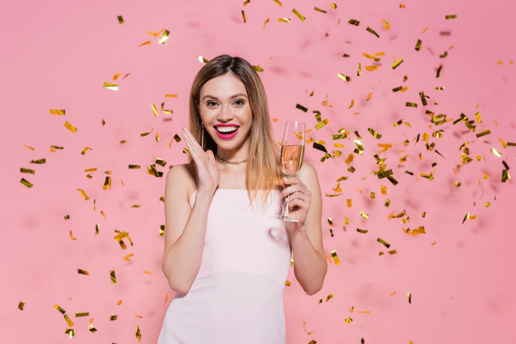 Ładna kobieta z szampanem oglądająca kamerę w pobliżu złotego konfetti podczas imprezy na różowym tle  - Zdjęcie, obraz