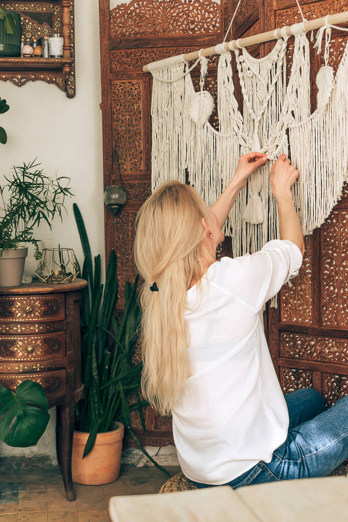 Une jeune femme blonde séduisante en chemisier blanc et en jean tisse des macrames.Le décor de la maison est accroché sur un écran en bois à motifs. L'intérieur de l'atelier est décoré avec des plantes d'intérieur.Vue arrière. - Photo, image