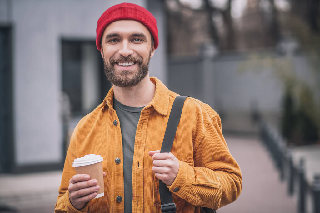 Kırmızı şapkalı ve elinde kahve fincanı olan turuncu ceketli bir adam. - Fotoğraf, Görsel