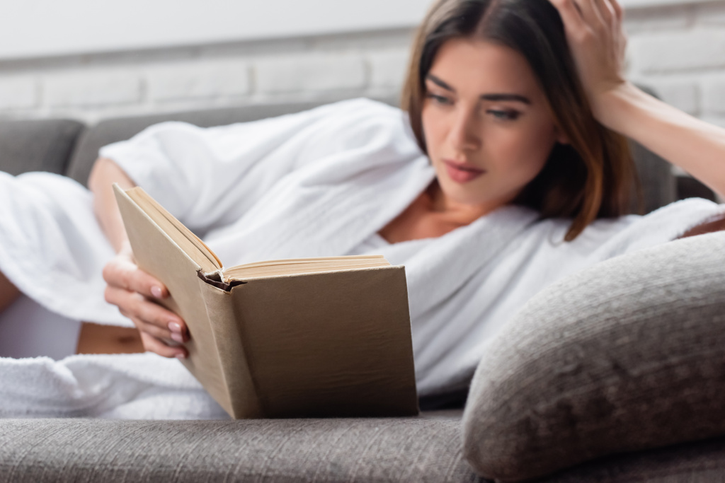 mujer adulta joven en el libro de lectura de albornoz en el sofá gris en casa - Foto, imagen