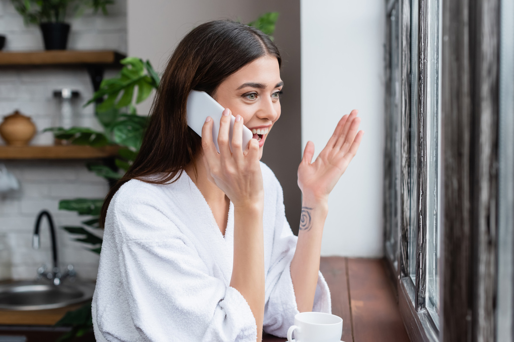 sonriente mujer adulta joven hablando por teléfono celular y haciendo un gesto con la mano cerca de la ventana en la sala de estar - Foto, imagen
