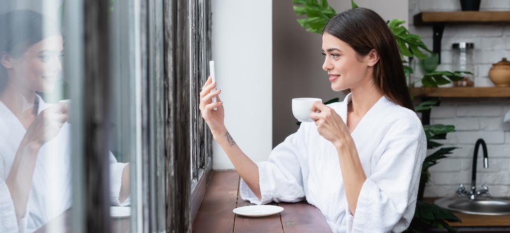 usmívající se mladá dospělá žena v županu se selfie na mobilu u okna v moderním podkroví, banner - Fotografie, Obrázek