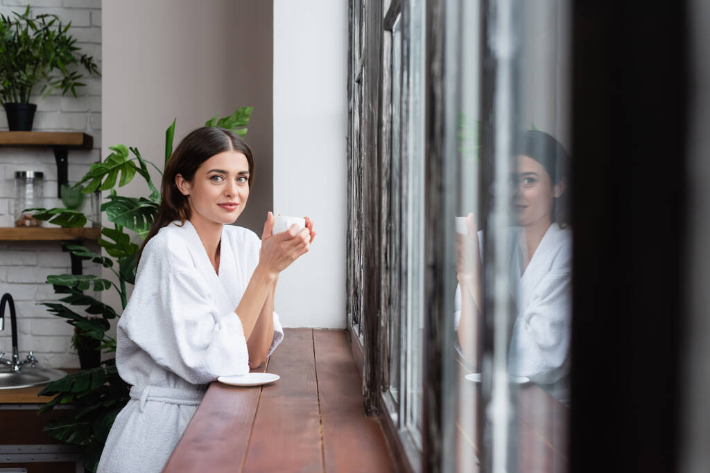 Modern oturma odasında yansıması olan mutlu, genç bir kadın pencerenin yanında bir fincan kahve tutuyor. - Fotoğraf, Görsel