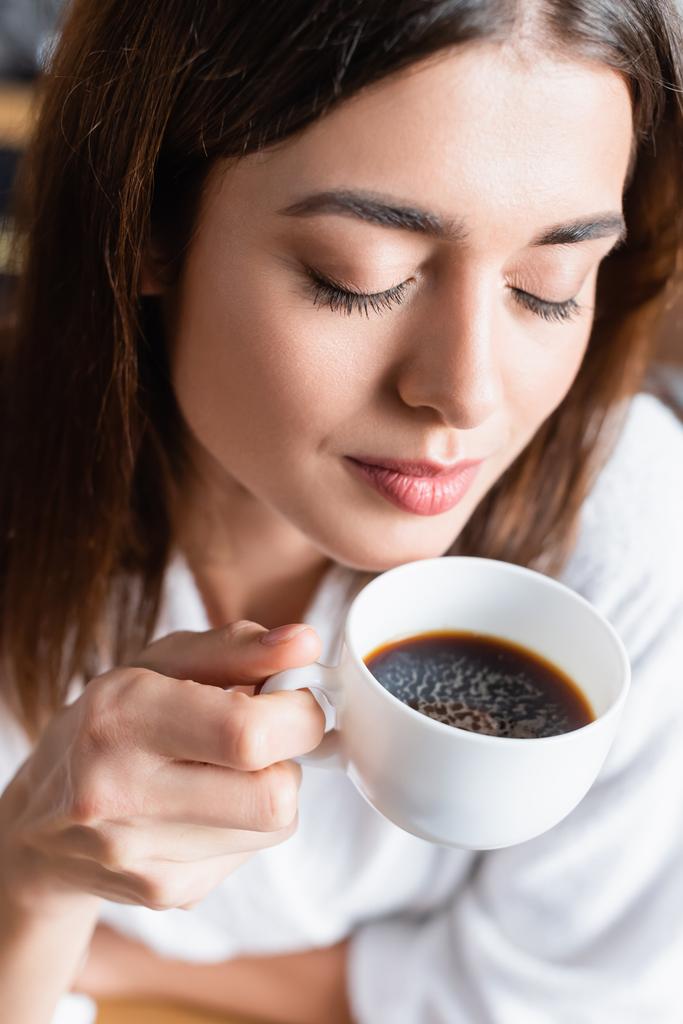 hoge hoek uitzicht op jonge volwassen vrouw drinken koffie met gesloten ogen op wazig achtergrond - Foto, afbeelding