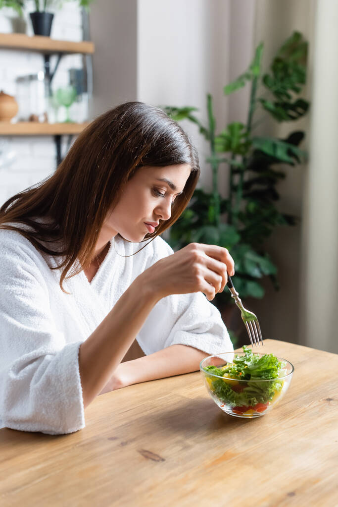 rozczarowana młoda kobieta w szlafroku jedząca sałatkę warzywną w nowoczesnej kuchni - Zdjęcie, obraz
