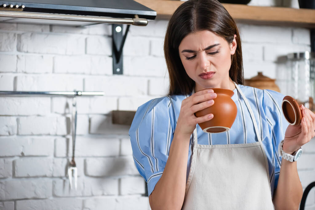 junge erwachsene Frau in Schürze blickt angewidert auf Tontopf in Küche - Foto, Bild