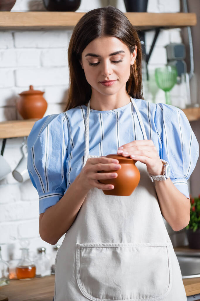 jovem mulher adulta alegre no avental que mantém o pote de barro em mãos na cozinha - Foto, Imagem