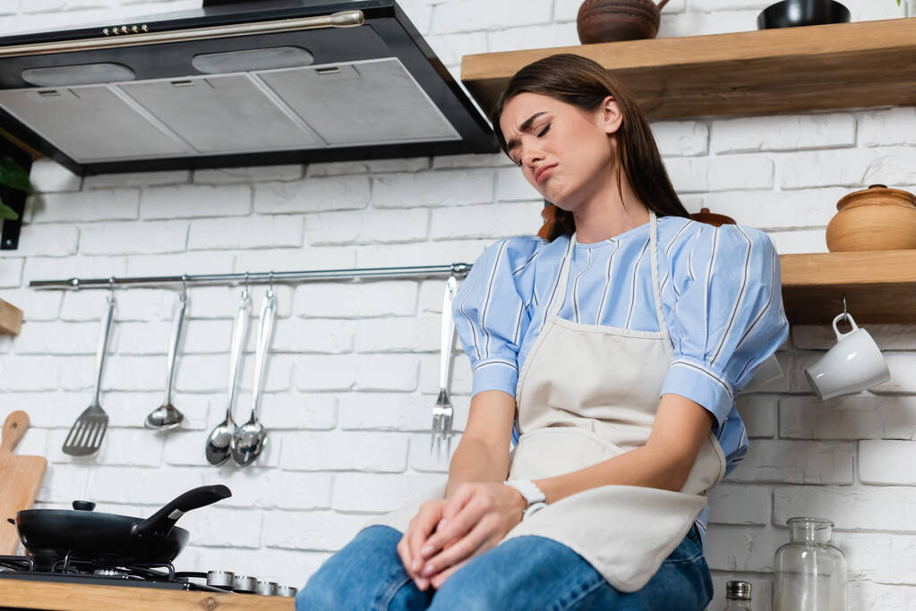 απογοητευμένη νεαρή ενήλικη γυναίκα κάθεται κοντά καυστήρα στην κουζίνα - Φωτογραφία, εικόνα