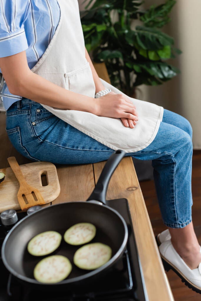 Önlüklü genç bir kadının modern mutfakta patlıcan dilimlerinin yanında oturması kısmi bir bakış açısı - Fotoğraf, Görsel