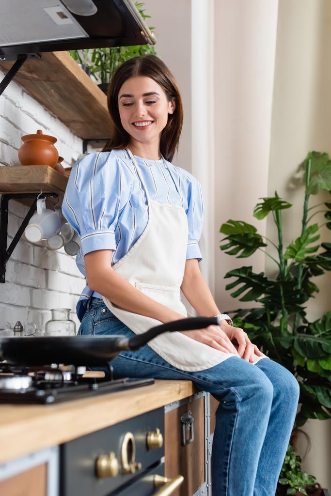 улыбающаяся молодая женщина в фартуке сидит рядом с сковородкой на современной кухне - Фото, изображение
