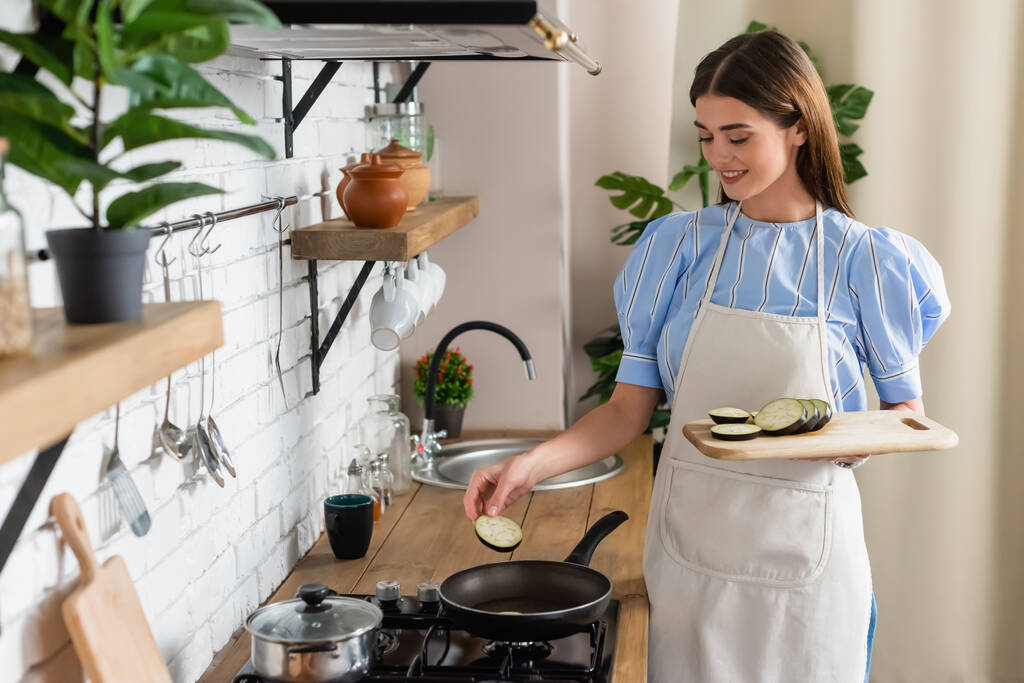 mujer adulta joven poniendo rebanadas de berenjena en la sartén en la cocina - Foto, imagen