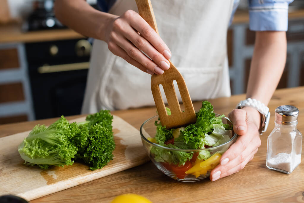 partial view of young adult woman mixing vegetables salad with spatula in kitchen - Photo, Image