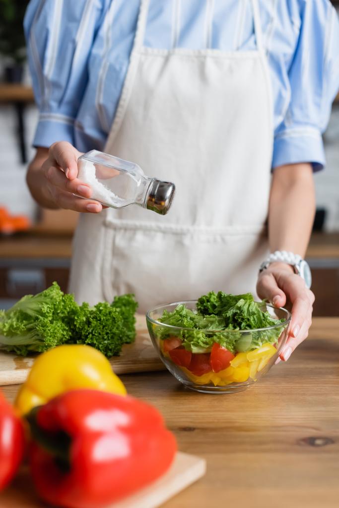 partial view of young adult woman in apron seasoning fresh vegetables salad with salt mill in modern kitchen - Photo, Image