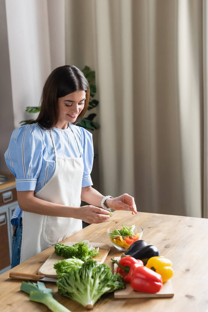 sonriente mujer joven adulta en delantal cocinar verduras frescas ensalada en la cocina - Foto, imagen