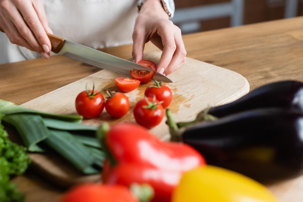 gedeeltelijk uitzicht op jonge volwassen vrouw snijden cherry tomaten op snijplank in moderne keuken - Foto, afbeelding