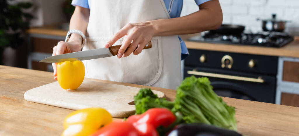 vue partielle de la jeune femme adulte coupant le poivron jaune sur la planche à découper dans la cuisine, bannière - Photo, image