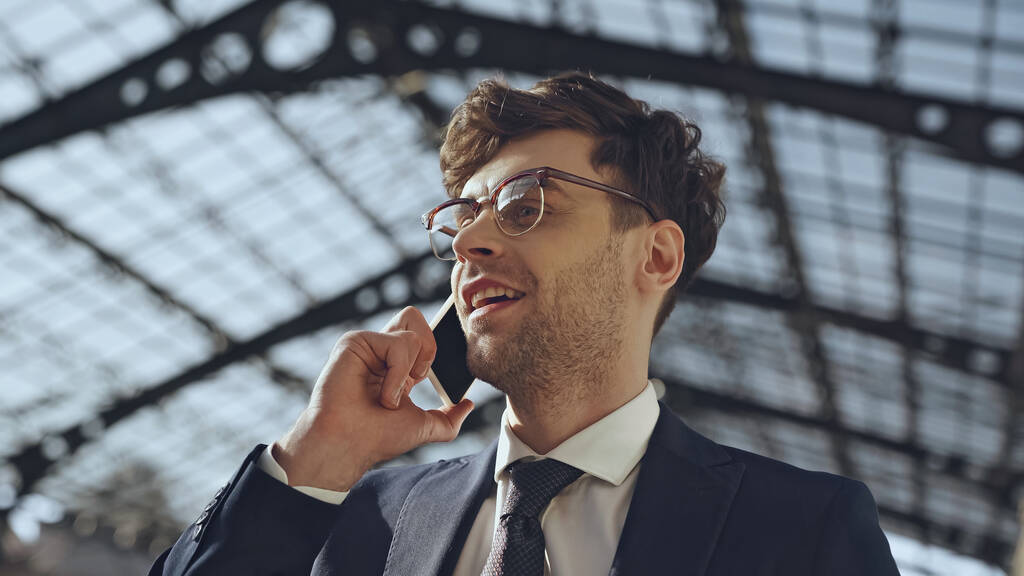 laag gezichtspunt van vrolijke zakenman in bril en pak praten op smartphone in modern winkelcentrum - Foto, afbeelding