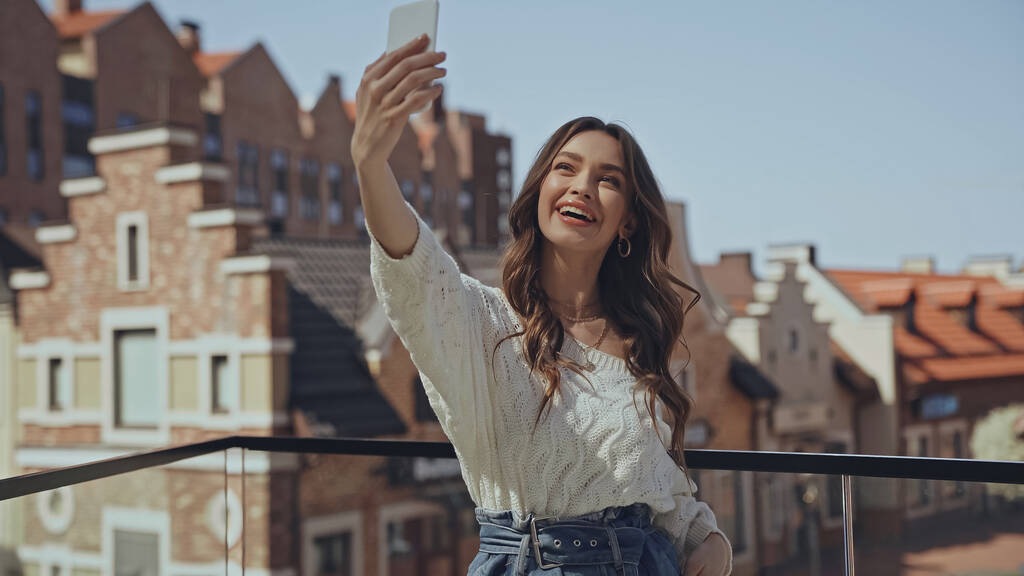 amazed young woman taking selfie near blurred buildings  - Photo, Image