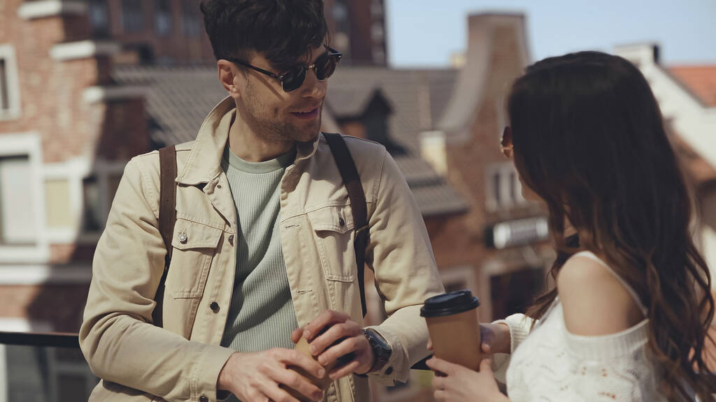стильная пара в солнечных очках, держащая бумажные стаканчики и разговаривая на улице  - Фото, изображение