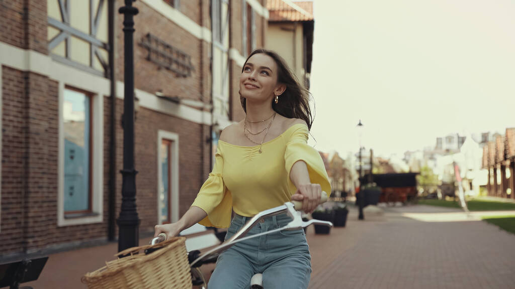 dromerige jonge vrouw in gele blouse glimlachen tijdens het rijden op de fiets buiten - Foto, afbeelding
