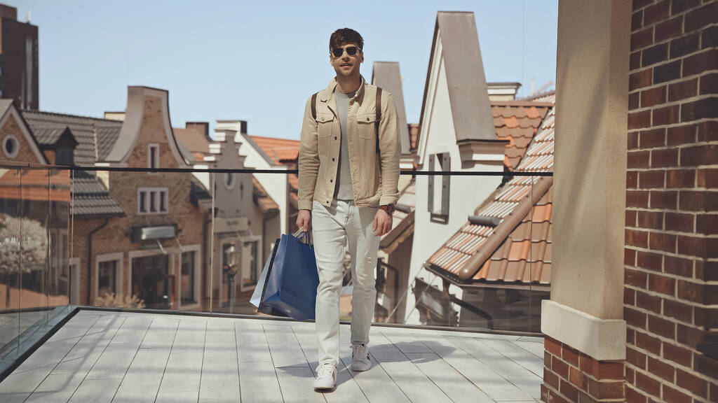 hombre con estilo en gafas de sol sosteniendo bolsas de compras y caminando fuera - Foto, imagen