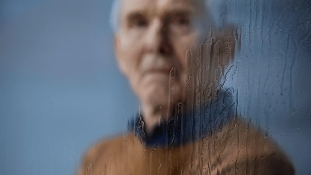 wazig portret van oudere man kijkend naar camera door regenachtig venster op grijze achtergrond - Foto, afbeelding