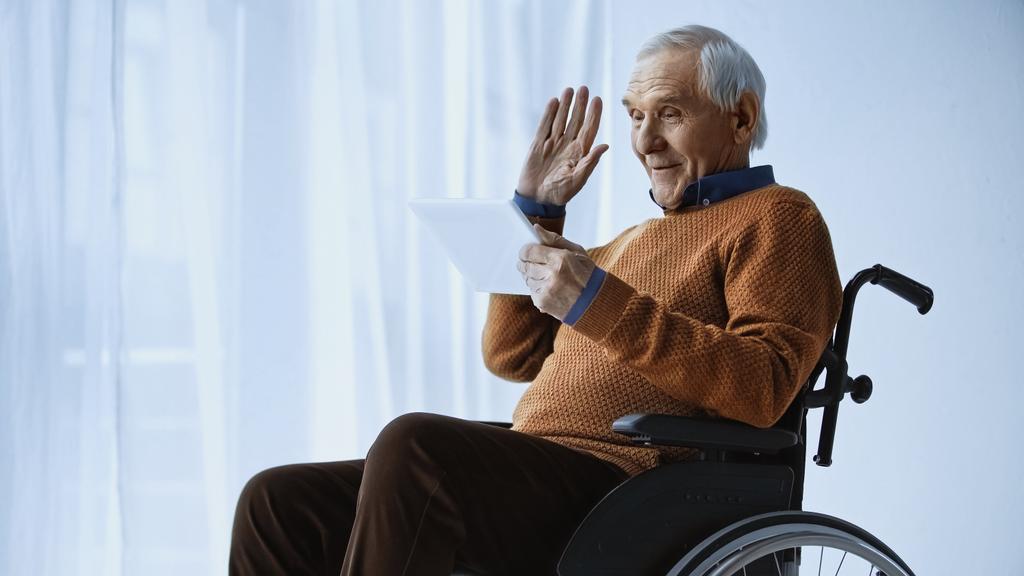 jubilado alegre en silla de ruedas saludando la mano y tener videollamada en la tableta en casa de retiro  - Foto, imagen