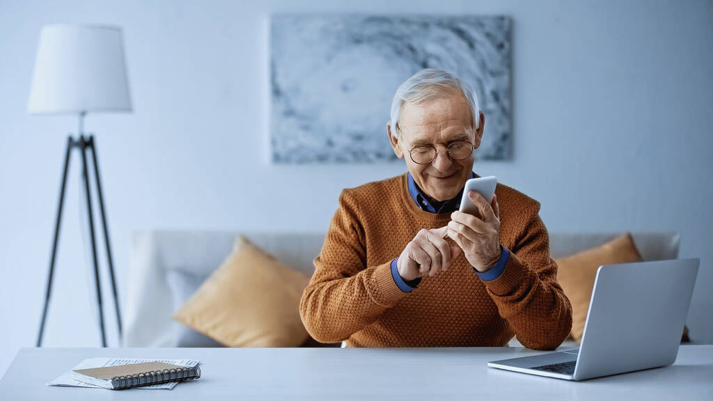 glimlachende oudere man zit in de buurt van laptop en berichten op smartphone thuis - Foto, afbeelding
