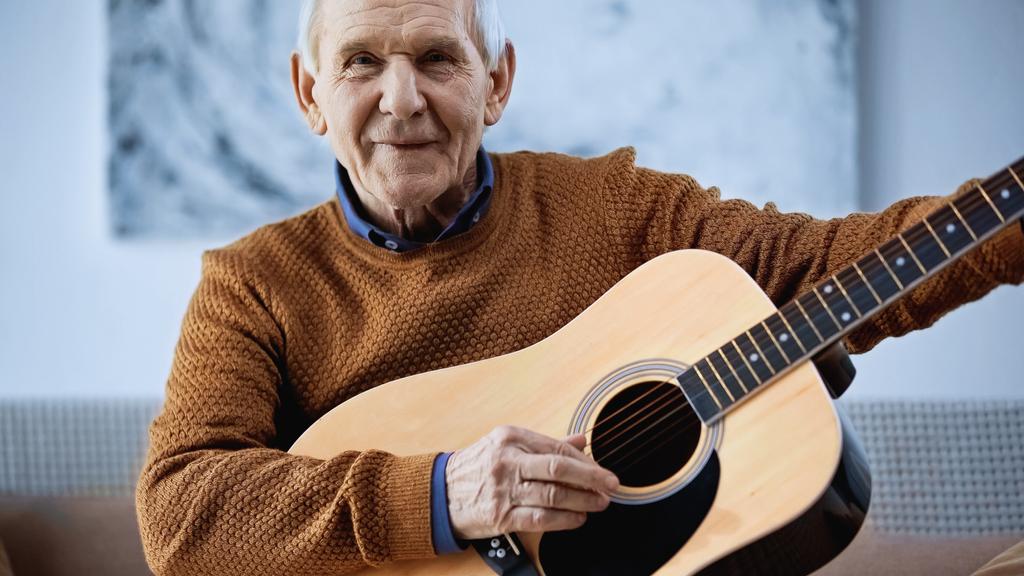 alegre anciano sentado en sofá con guitarra en la sala de estar  - Foto, imagen