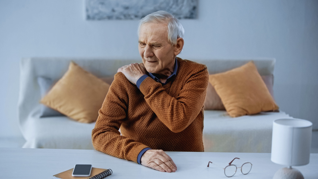 trpící starší muž sedí u stolu a dotýká se svého ramene doma - Fotografie, Obrázek