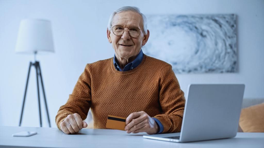 gelukkige oudere man met een bril in de buurt van laptop en mobiele telefoon en met een creditcard in de woonkamer - Foto, afbeelding