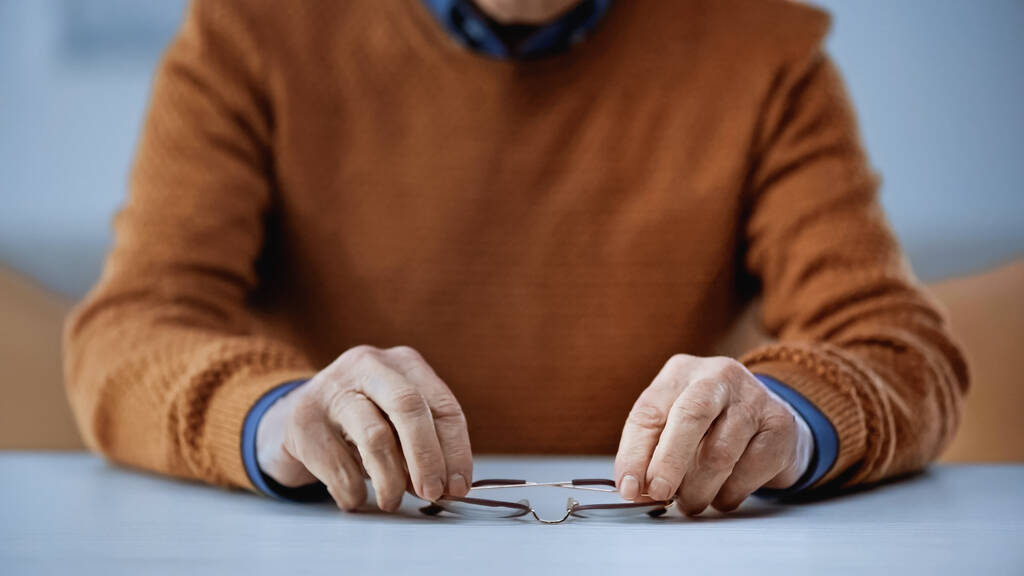 vista cortada de homem idoso segurando óculos em mãos sobre fundo cinza e bege - Foto, Imagem