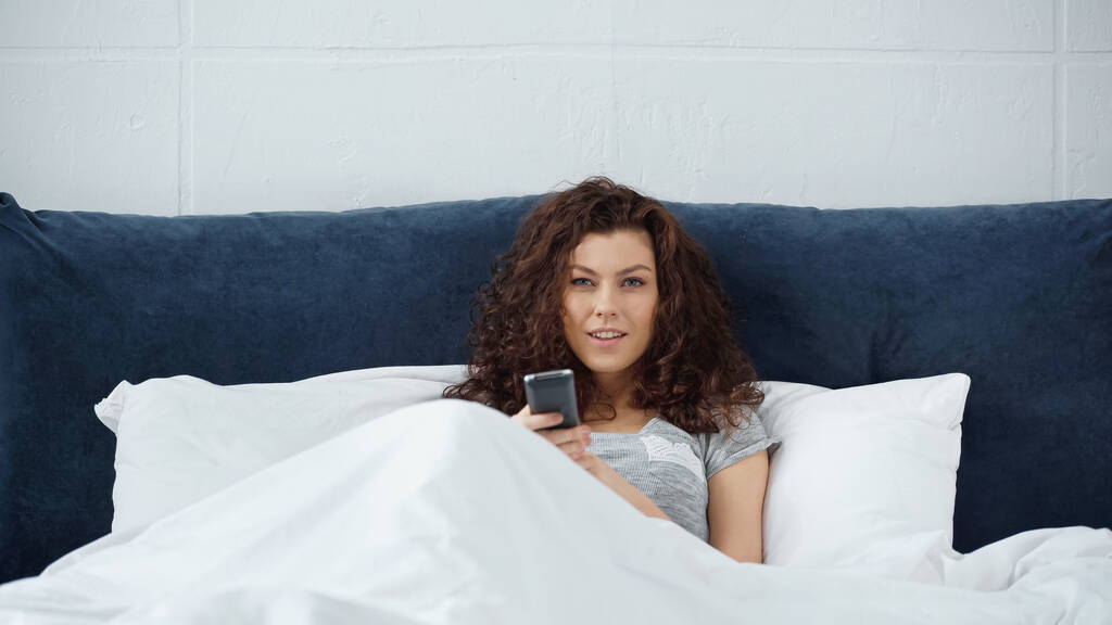 happy young woman holding remote controller in bed  - Photo, Image