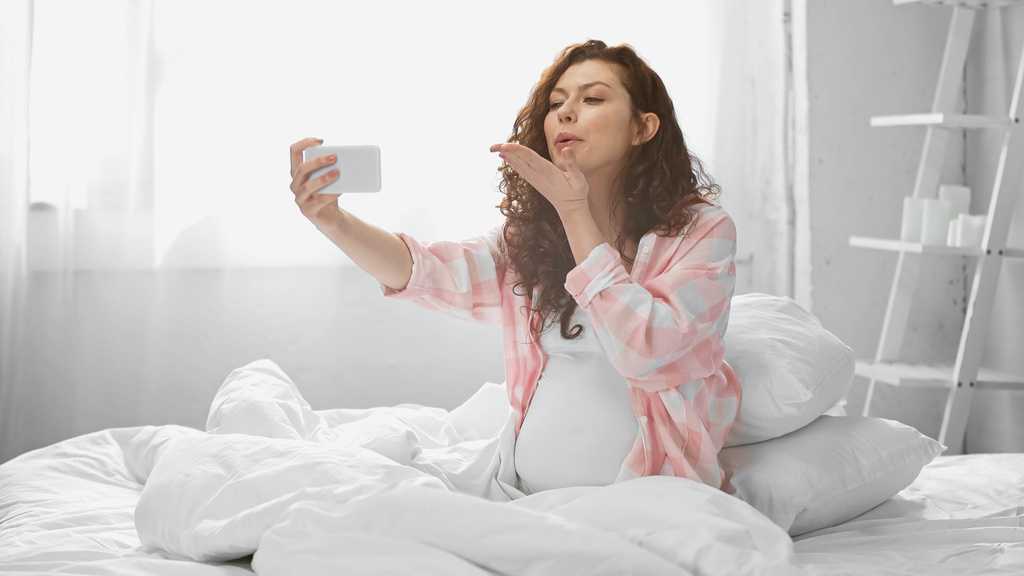 lockige und schwangere Frau verschickt Luftkuss, während sie ein Selfie im Schlafzimmer macht  - Foto, Bild