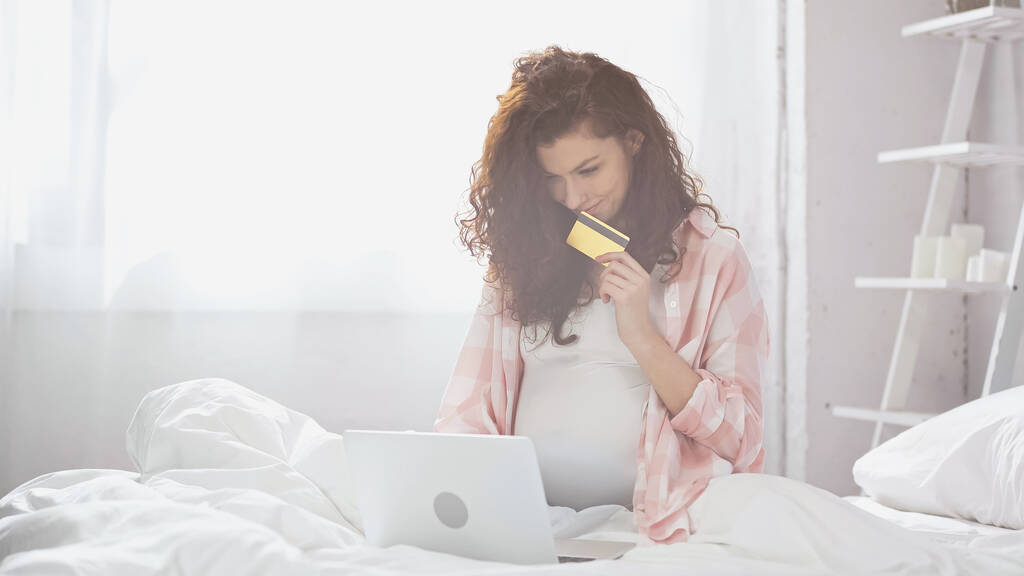 счастливая беременная молодая женщина с кредитной картой во время использования ноутбука в постели  - Фото, изображение