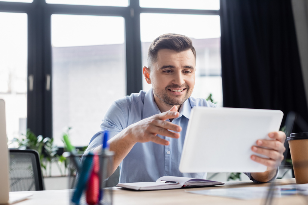 Usmívající se podnikatel pomocí rozmazané digitální tablet v blízkosti notebooku a kávy jít  - Fotografie, Obrázek