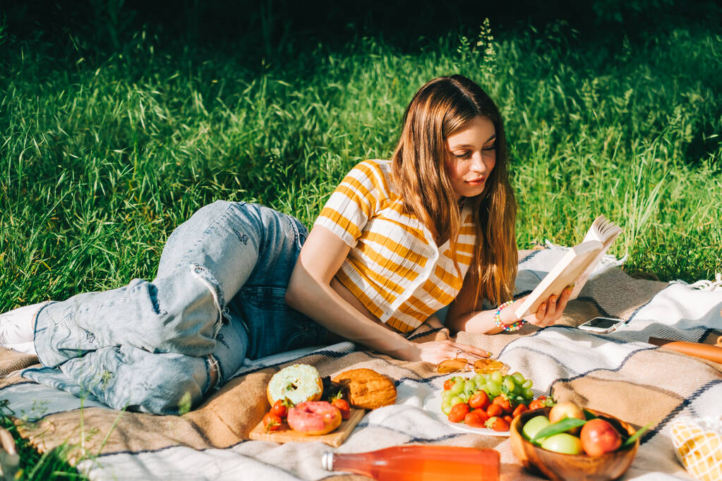 Nuori nainen lukee kirjaa ulkona, luonnonpuistossa piknikillä, makaa piknikpeitolla ja nauttii kesäsäästä. - Valokuva, kuva