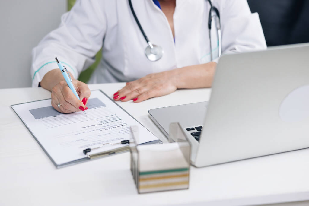 Arzt im weißen Kittel schreibt Notizen mit Laptop am Schreibtisch - Foto, Bild
